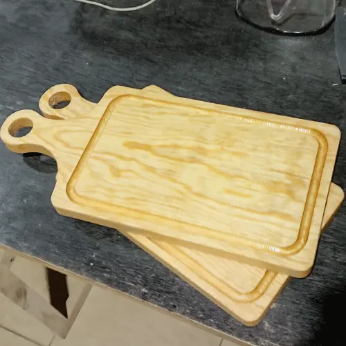 Dos tablas rectangulares de pino con mango apiladas sobre un banco de trabajo oscuro en un taller.