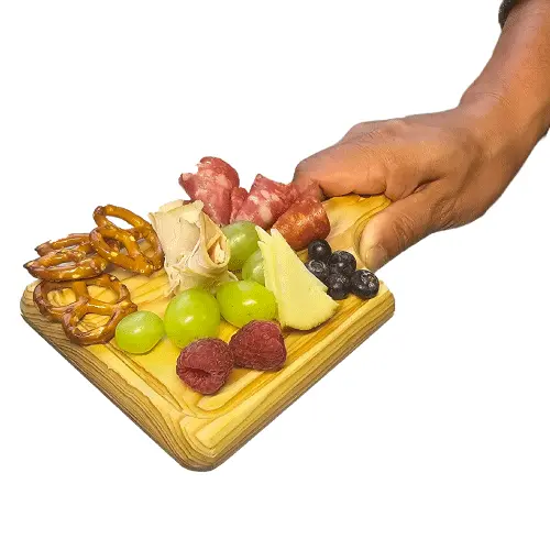 mini-tabla-pino-charcuteria-porciones-individuales.webp Mano de una persona sosteniendo una mini tabla cuadrada de madera de pino servida con pretzels, queso, uvas, frambuesas y carnes frías.
