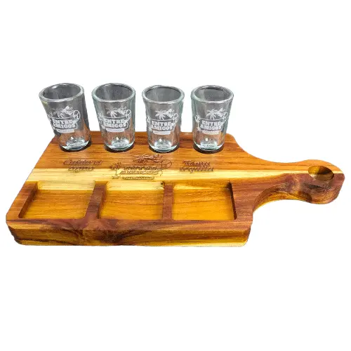 Tabla de madera con mango para 4 caballitos de tequila, mostrando los vasos grabados y el contraste de vetas claras y oscuras de la madera.