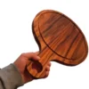 Mano de una persona sosteniendo firmemente en el aire una gruesa tabla redonda de madera de teca por el mango.