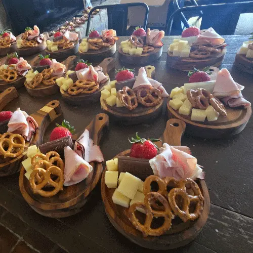catering-mini-tablas-charcuteria-eventos-perspectiva.webp Mesa larga llena de mini tablas de madera circulares servidas con porciones individuales de carnes frías, queso, fresas y pretzels para un evento.