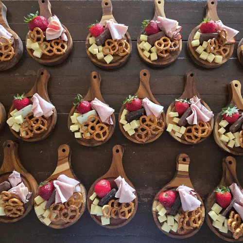 catering-mini-tablas-charcuteria-eventos-perspectiva2.webp Vista superior de múltiples mini tablas de madera circulares ordenadas en filas, listas con aperitivos de charcutería, fresas y pretzels.