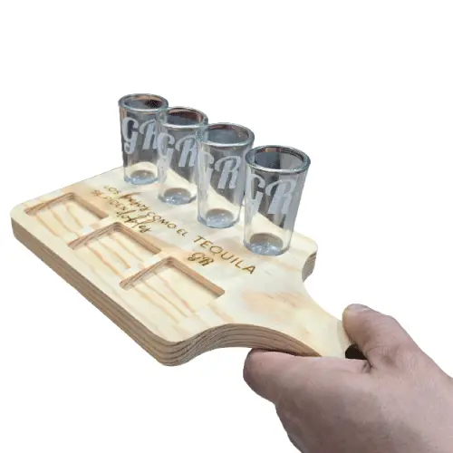 Mano sosteniendo una tabla tequilera de madera personalizada de Santos Customs Designs, mostrando el grabado láser "Los besos como el tequila se piden dobles", 4 caballitos de vidrio grabados con iniciales GR y compartimentos para botanas.