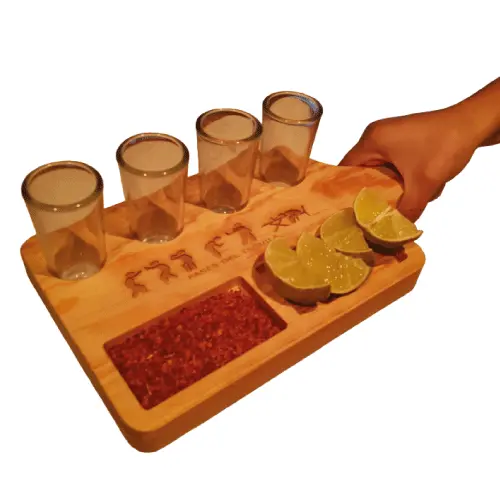 Mano sosteniendo una tabla de madera para tequila de Santos Customs Designs, equipada con 4 caballitos de vidrio, chile, limón y un grabado de las Fases del Tequila.