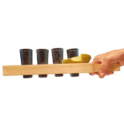 Mano sosteniendo una tabla tequilera de madera (tipo paleta) de Santos Customs Designs con cuatro caballitos oscuros y rodajas de limón, aislada sobre fondo blanco.